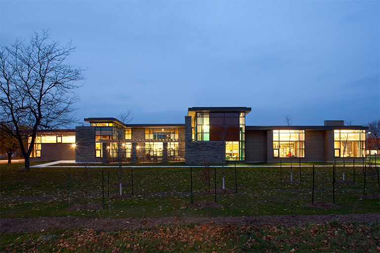 South-facing view of a modern library with warm interior lighting, large windows, and a mix of brick and stone cladding at dusk.